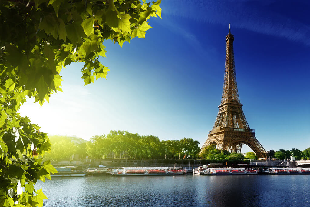 picture of Eiffel Tower on a bright blue sky day with scene river in the foreground with ferries in it.